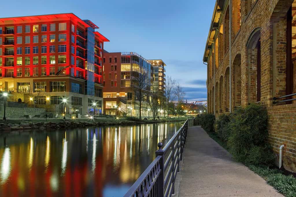 Attrazioni serali lungo il Reedy Riverwalk nel centro di Greenville, nella Carolina del Sud, mentre le luci brillano e si riflettono sull'acqua.