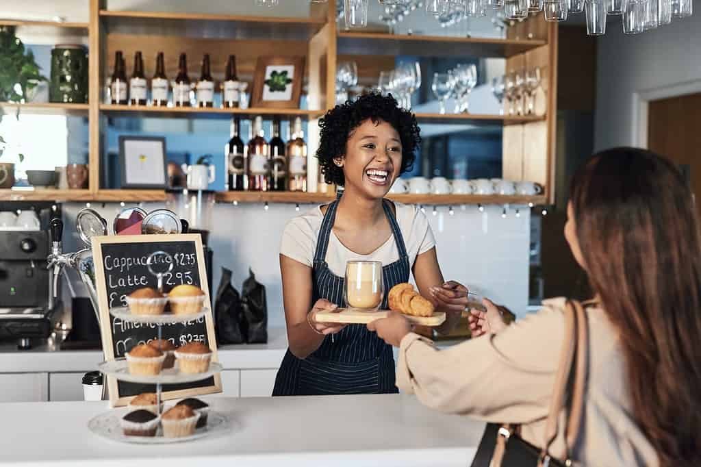 Donna felice, barista e cliente al servizio del bar per servizio, pagamento o ordine al bancone del bar.  Persona africana, cameriera o dipendente in un ristorante di piccole imprese che aiuta il cliente alla cassa