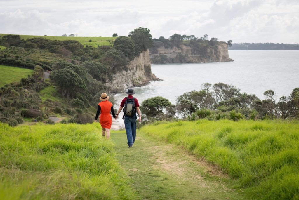Coppia che si tiene per mano, camminando lungo la passeggiata costiera di Long Bay.  Auckland.