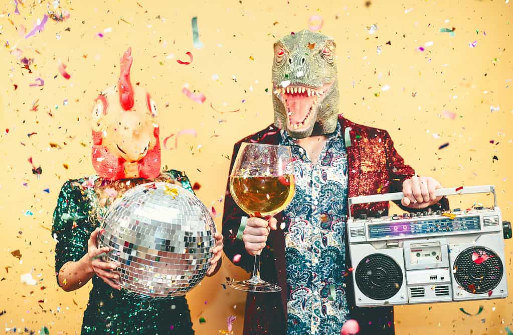 Coppia pazza che festeggia il capodanno indossando una maschera da pollo e dinosauro t-rex - Giovani alla moda che si divertono bevendo champagne e ascoltando musica con boombox vintage - Assurdo e concetto di vacanze