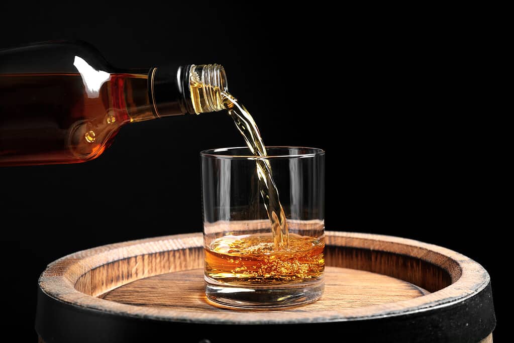 Versare il whisky dalla bottiglia in vetro su botte di legno su sfondo nero, primo piano