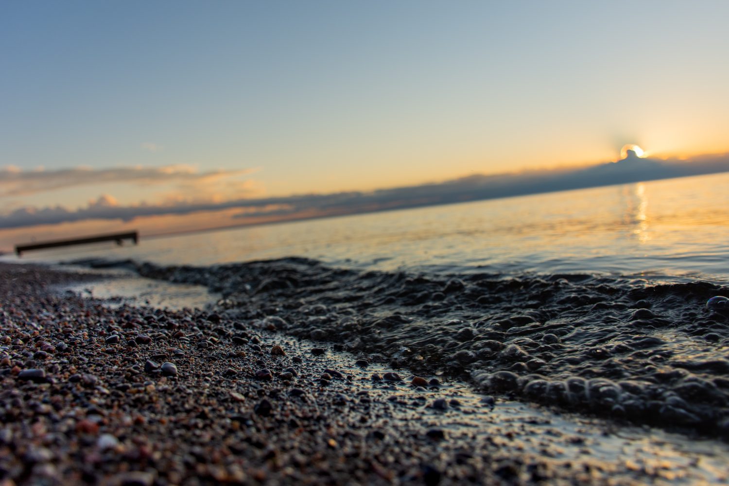 Primo piano su rocce di sabbia e piccole onde con messa a fuoco morbida sul molo di legno e sole nascente che penetra attraverso nuvole scure sopra l'orizzonte e il lago Saint-Jean, Quebec, Canada, in una calda mattina estiva