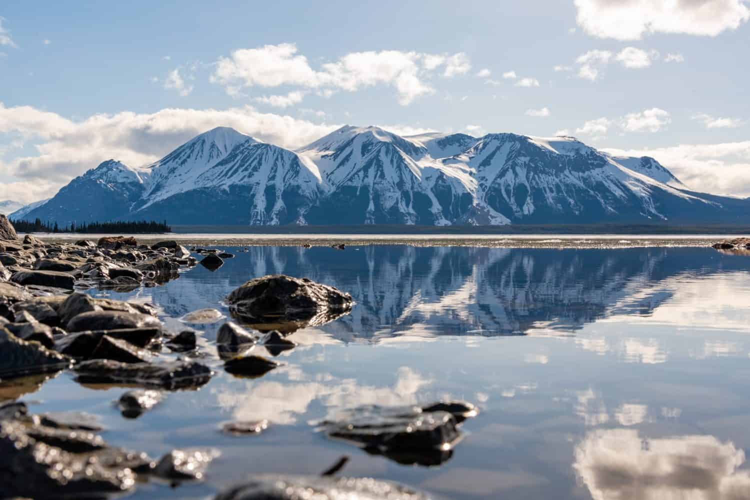 Splendida vista sui Monti Atlin nella Columbia Britannica settentrionale durante la primavera in una perfetta giornata di cielo azzurro con nuvole e riflessi nell'acqua calma sottostante.  Scenico, vista per casa, ufficio art. 