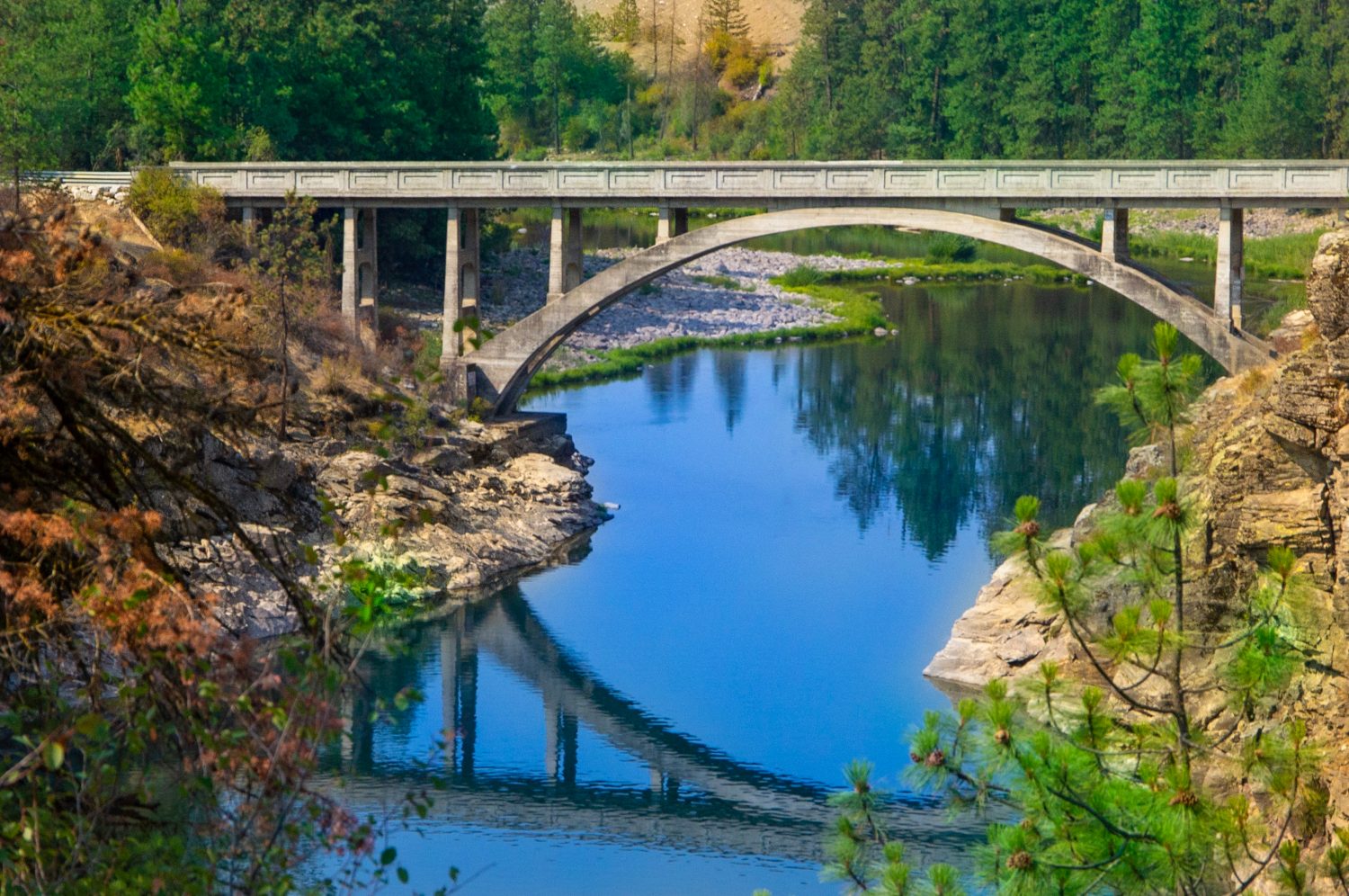 Post Falls, ID: 6 agosto 2018;  Il ponte ad arco in cemento attraversa il fiume Spokane con il cielo azzurro e l'architettura che si riflette sull'acqua in esposizione temporale con alberi sempreverdi sullo sfondo.