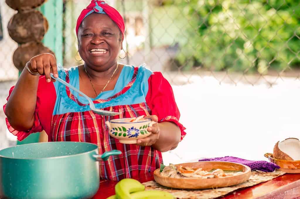 Una bella donna nera di etnia Garífuna sorride alla telecamera mentre mette la zuppa calda nel piatto.