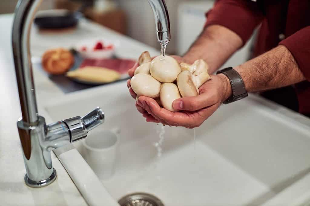 L'uomo sconosciuto tiene in mano un fungo fresco e lo pulisce sotto uno spruzzo nel lavello della cucina.