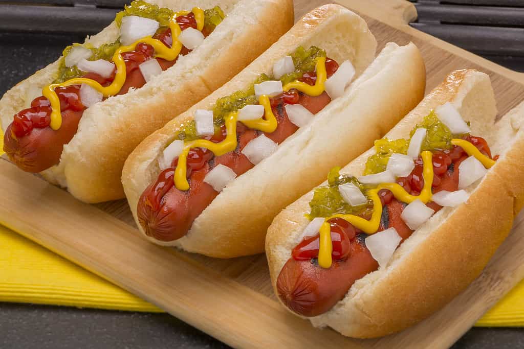 Tre hot dog con senape, ketchup, sottaceti e cipolle su un tagliere di legno