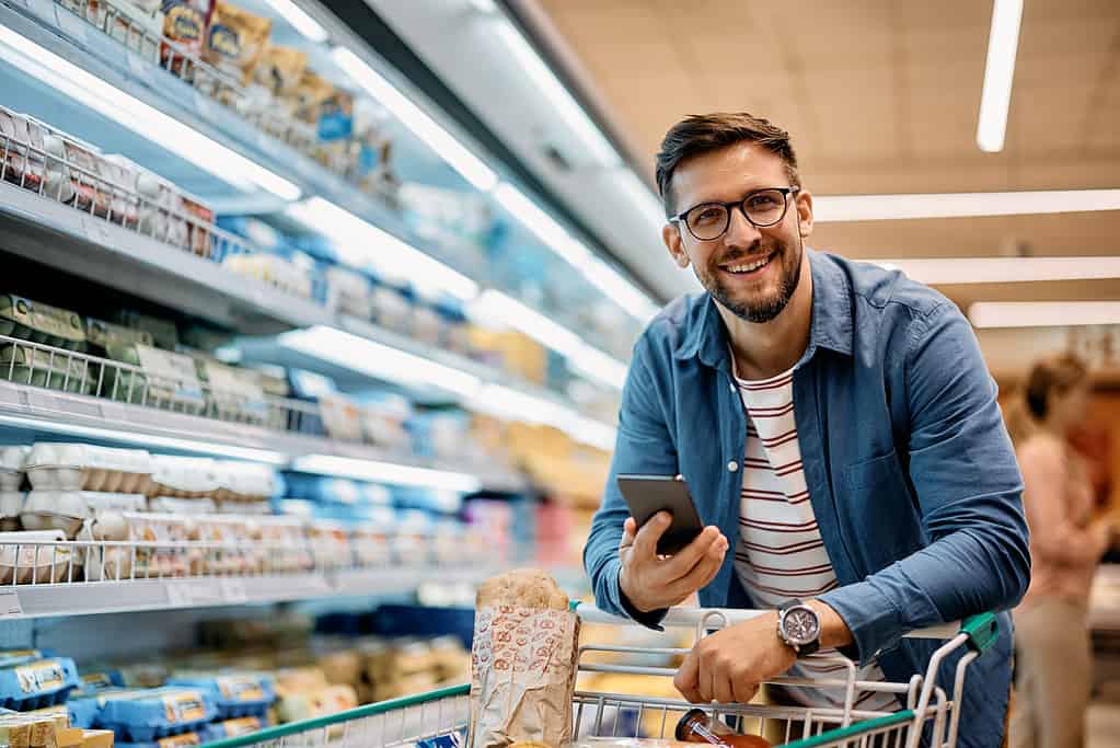 Uomo felice che utilizza l'app del telefono cellulare mentre acquista generi alimentari al supermercato e guarda la fotocamera.