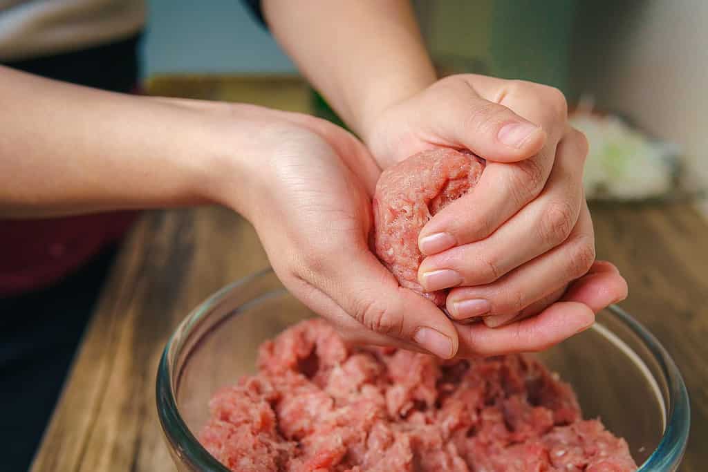 Primo piano delle mani di una donna che prepara la carne macinata per fare gli hamburger, la carne è ancora cruda e sta aggiungendo gli ingredienti, bella atmosfera in cucina.