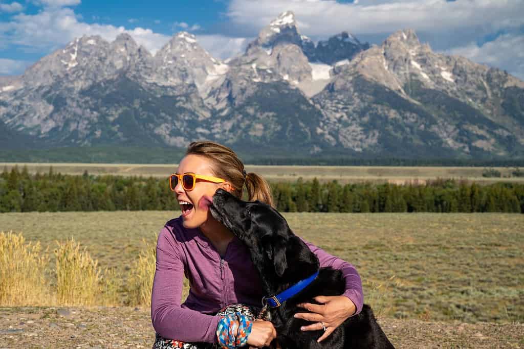 Una donna abbraccia e tiene in braccio il suo cane labrador retriever nero di fronte alle montagne del Parco Nazionale Grand Teton a Jackson, nel Wyoming, mentre il cane le lecca il viso