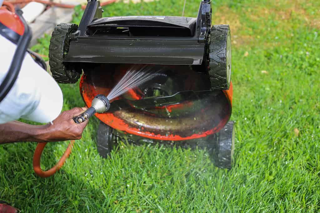 Un uomo afro-americano nero che spruzza e pulisce un tosaerba con un tubo dell'acqua all'esterno in estate