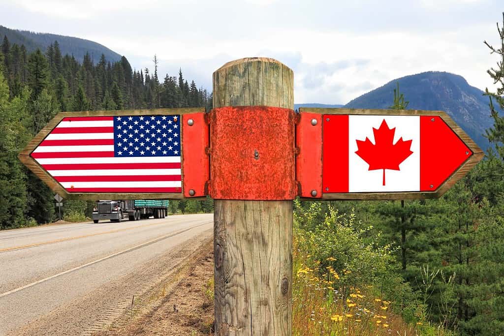 Cartello in legno con due frecce: bandiere nazionali americane e canadesi sulla strada statale con paesaggio naturale sullo sfondo.  Segnale di direzione in movimento del Canada e dell'America