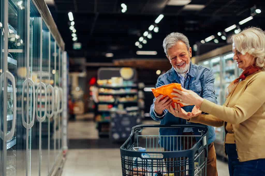 Coppia di anziani che sceglie la spesa in un supermercato, tenendo in mano una borsa di prodotto irriconoscibile e leggendo l'etichetta sul retro della confezione
