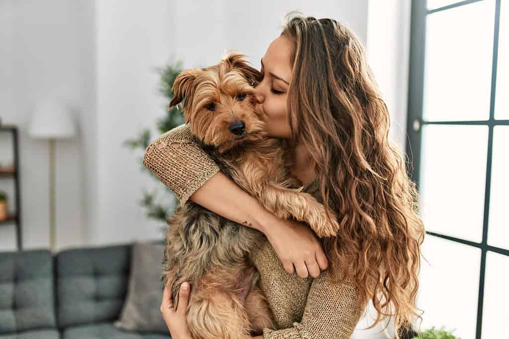 Giovane bella donna ispanica che bacia e abbraccia il cane in piedi a casa