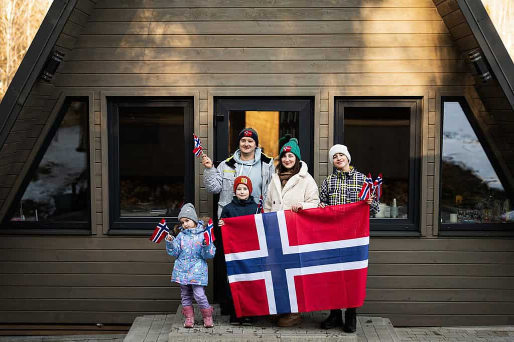 Ritratto di famiglia con bambini fuori dalla baita che tengono bandiere norvegesi. Cultura scandinava, popolo norvegese.
