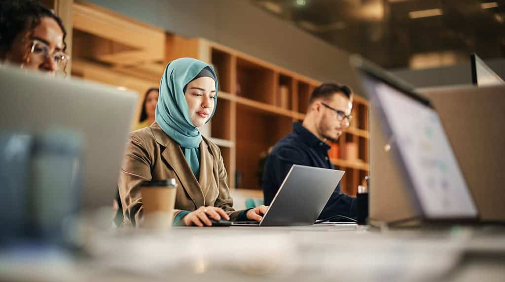 Ritratto di donna musulmana concentrata che usa il portatile in un ufficio spazioso e moderno. Account Specialist sorridente mentre trascorre una giornata produttiva. Colleghi attivi che lavorano sui computer sullo sfondo.