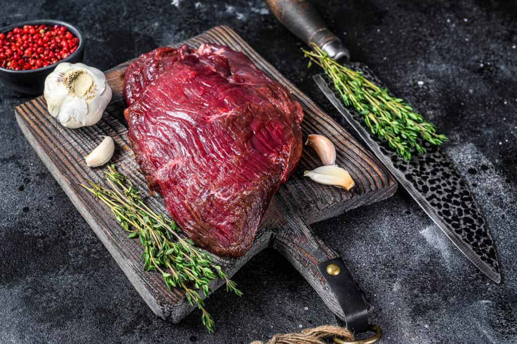 Carne di cervo cruda su un tagliere con erbe aromatiche. Sfondo nero. Vista dall'alto