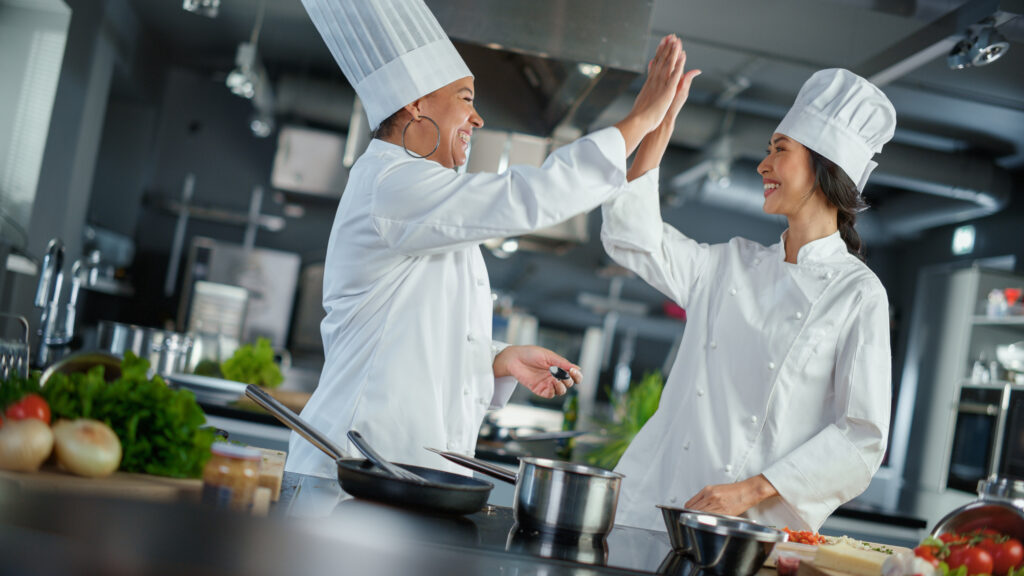 Cucina del ristorante: Ritratto di chef asiatiche e nere che preparano piatti, assaggiano cibo, si danno il cinque in una festa di successo. Due professioniste cucinano cibo delizioso e autentico, pasti sani